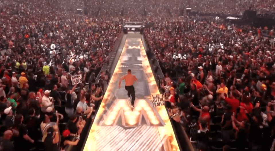 A wrestler running down a long ramp toward the ring, with a huge crowd cheering on either side.