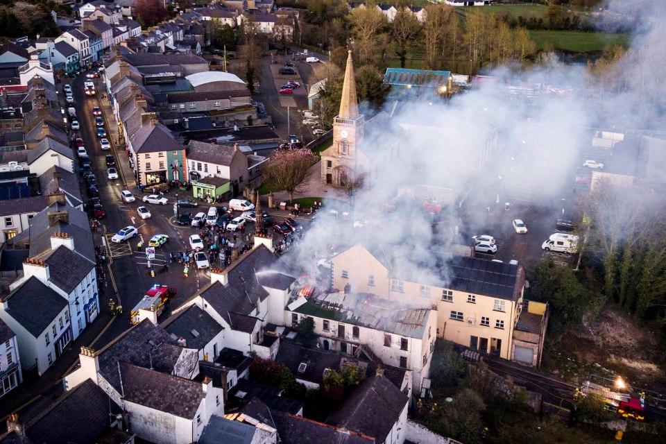 Aerial view of a town with smoke rising from a burning building, surrounded by emergency vehicles and onlookers.