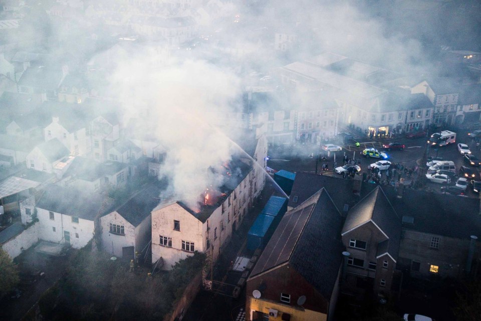 Aerial view of a town where a building is on fire, emitting a large cloud of smoke, with emergency services and onlookers on the streets.