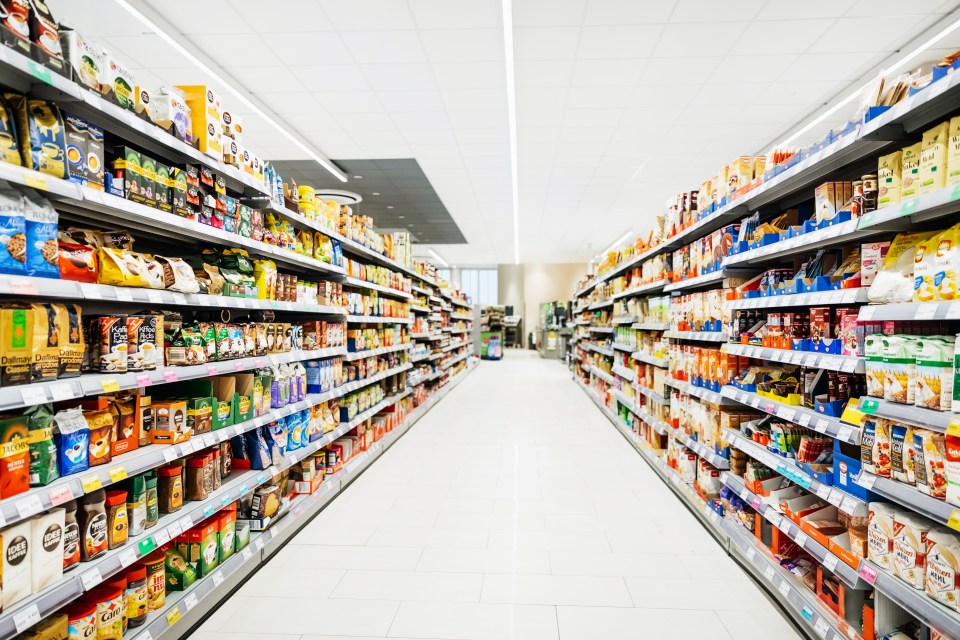 A brightly lit supermarket aisle with shelves full of products on both sides, including packaged coffee, flour, and other groceries.