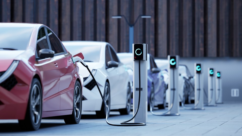 Illustration of a red electric car charging at a station on a city street, with other electric cars and charging stations in the background.