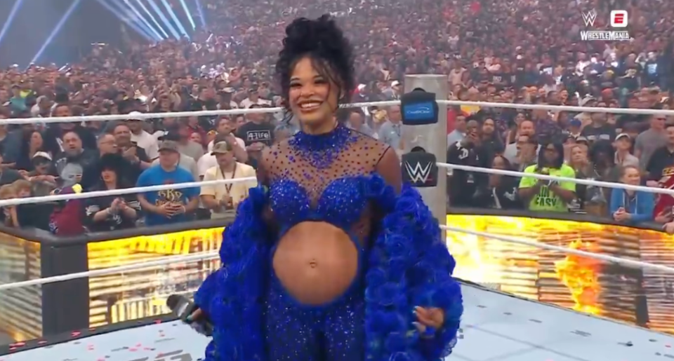 A smiling pregnant woman in a royal blue sequined jumpsuit with ruffled sleeves and a cut-out over her belly, stands in a wrestling ring before a cheering crowd.