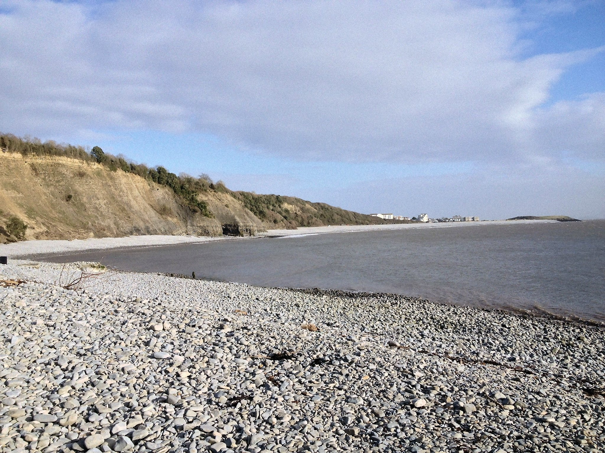 A wide view of a pebble beach next to the sea, with a rocky cliff covered in trees to the left, and distant buildings on the right horizon.
