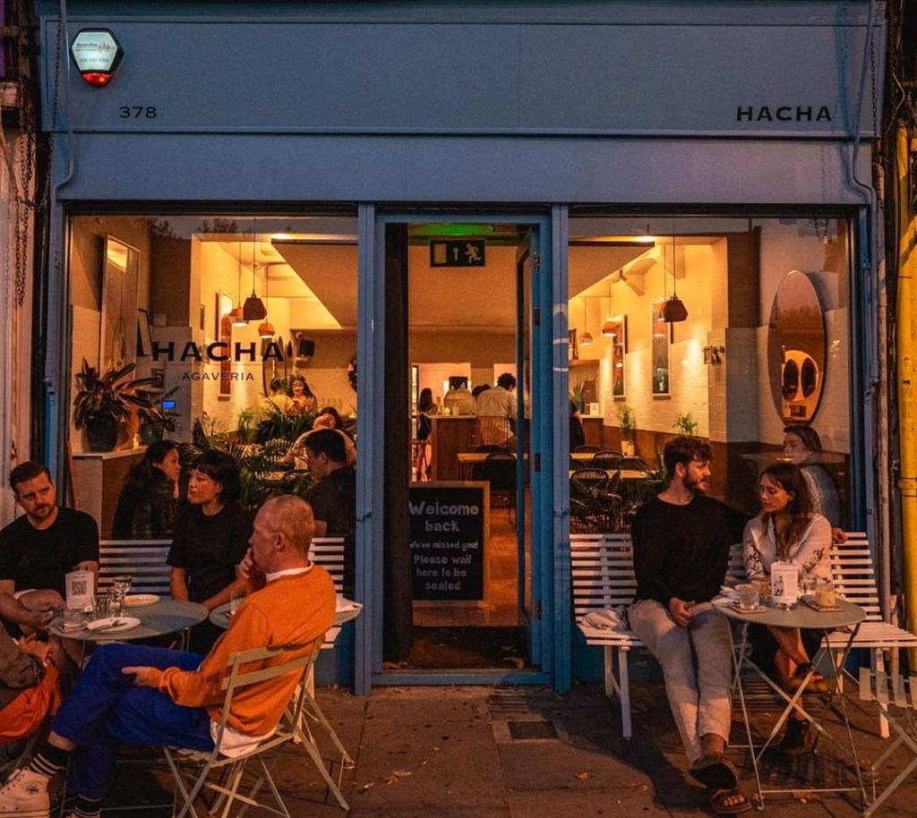 People seated at outdoor tables and inside Hacha Agavera, a bar with a light blue exterior.