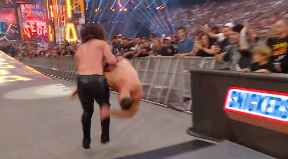 A male wrestler in black attire throwing another male wrestler over the barrier into the crowd.