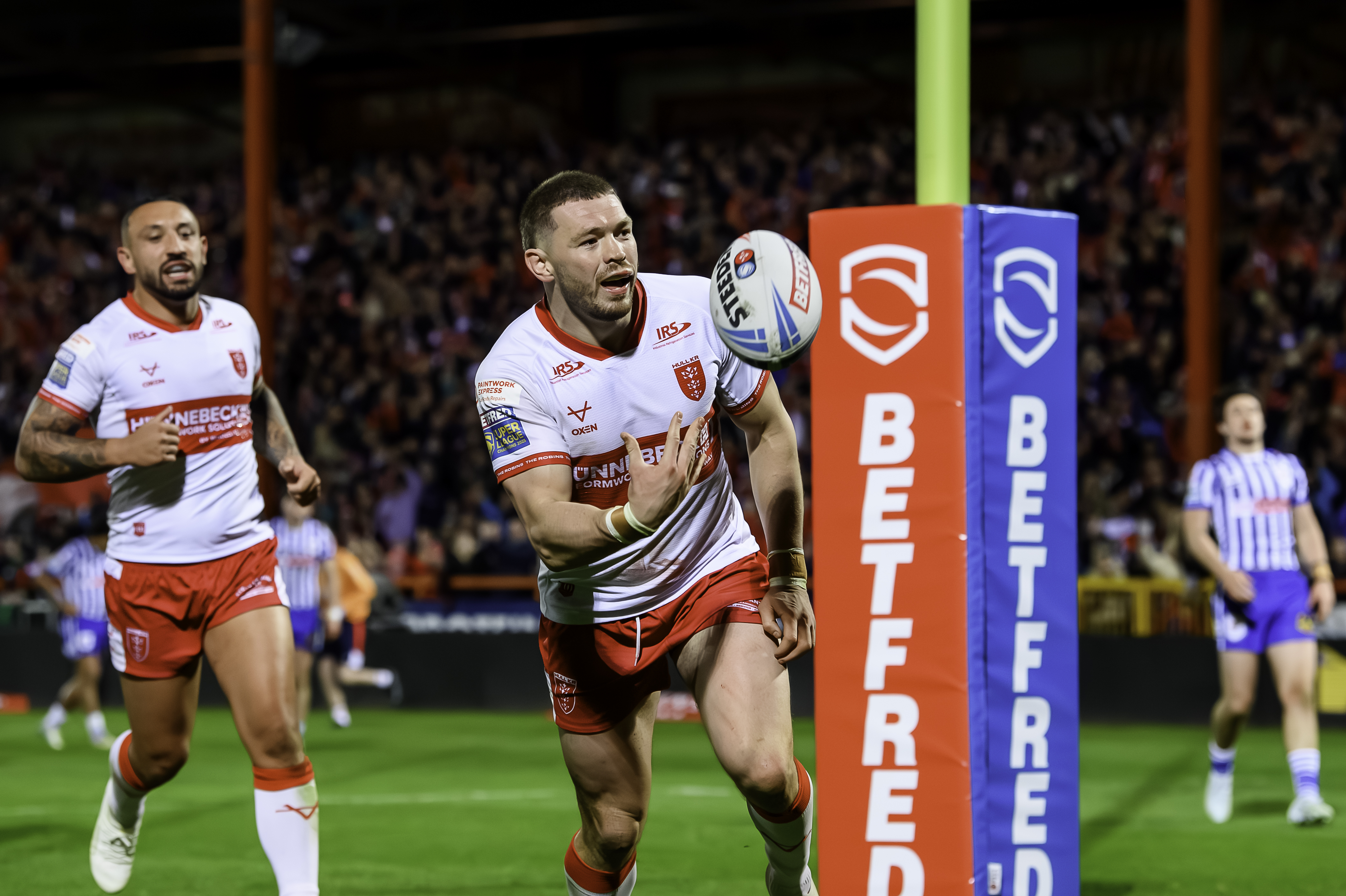 A rugby player in a white and red uniform scoring a try, with a teammate celebrating behind him.