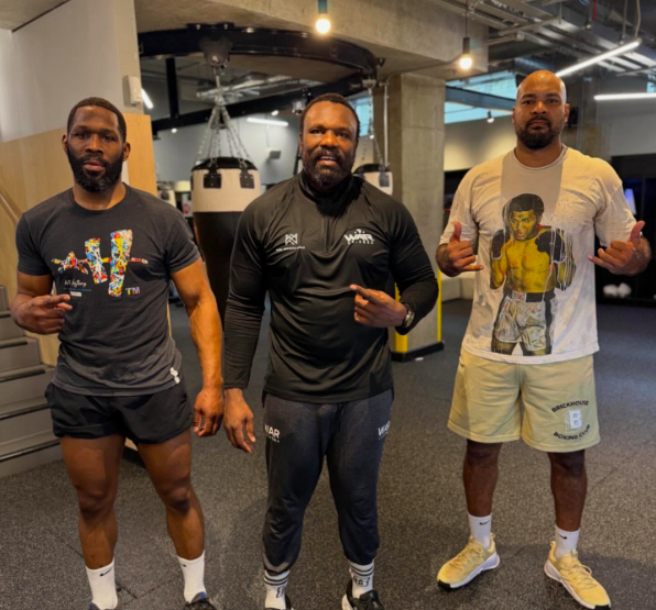 Three men in a gym, two pointing at the man in the middle who is wearing a black long-sleeved shirt with "WAR Sports" logo.