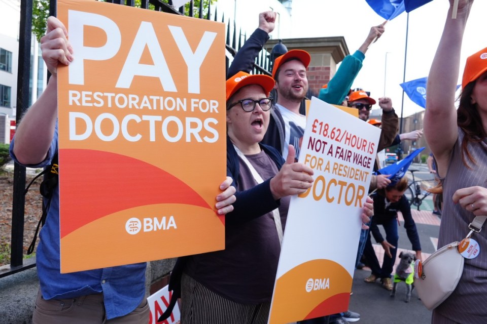 Protesters at an industrial action hold signs demanding pay restoration for doctors and stating that £18.62/hour is not a fair wage for a resident doctor.
