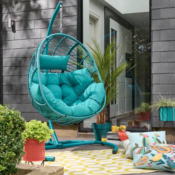 A turquoise hanging egg chair with turquoise cushions on a patio with a yellow and white rug.