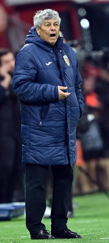A soccer coach in a blue winter coat with a team patch, gray hair, and an open mouth, stands on the edge of a soccer field with a player in a yellow jersey partially visible on the left.