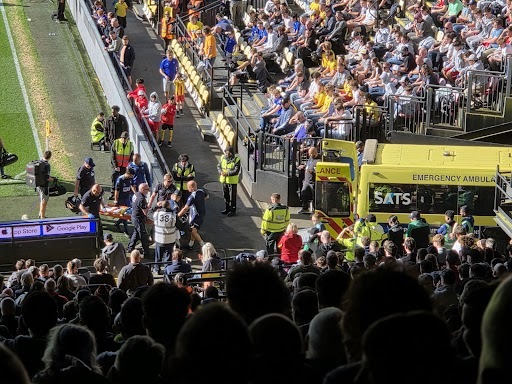 Emergency responders attending to an injured person on a stretcher next to an ambulance on a soccer field, surrounded by spectators.