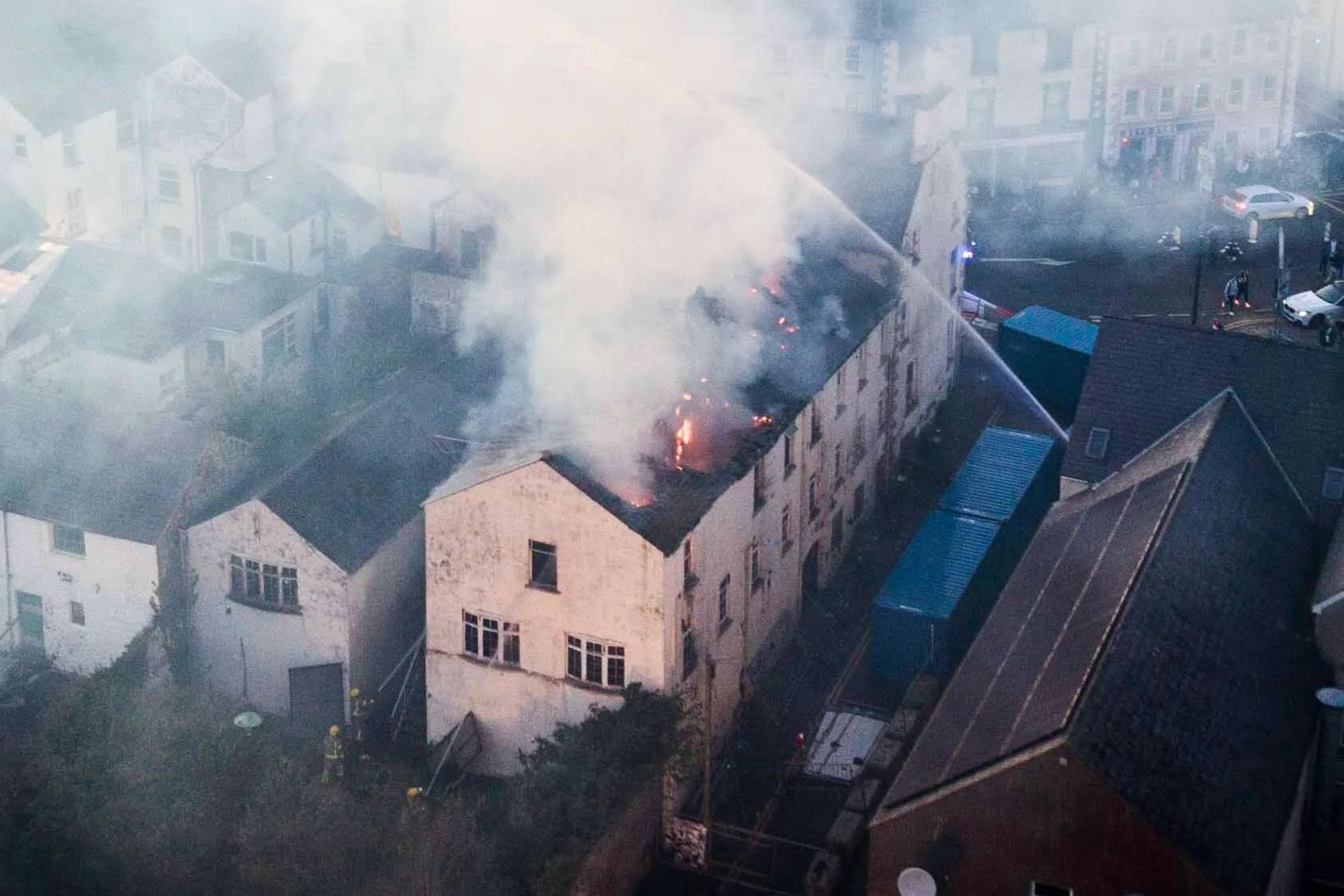 An image collage containing 1 images, Image 1 shows Aerial view of a town where a building is on fire, emitting a large cloud of smoke, with emergency services and onlookers on the streets