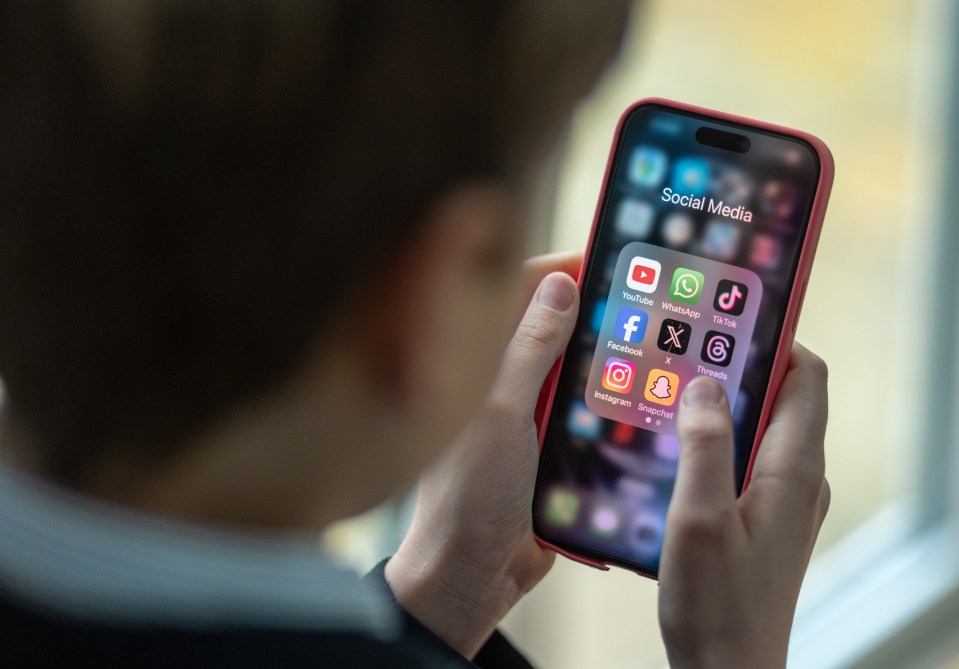 A 12-year-old boy in a school uniform holds a red iPhone displaying a folder of social media apps, including YouTube, WhatsApp, TikTok, Facebook, X, Threads, Instagram, and Snapchat.