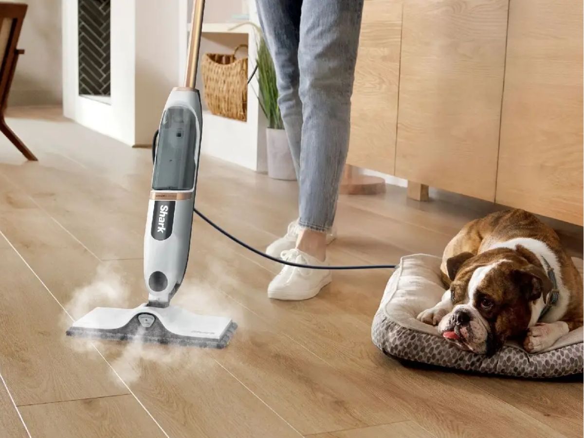 A person uses a Shark steam mop on a hardwood floor, while a bulldog lies on a dog bed nearby.