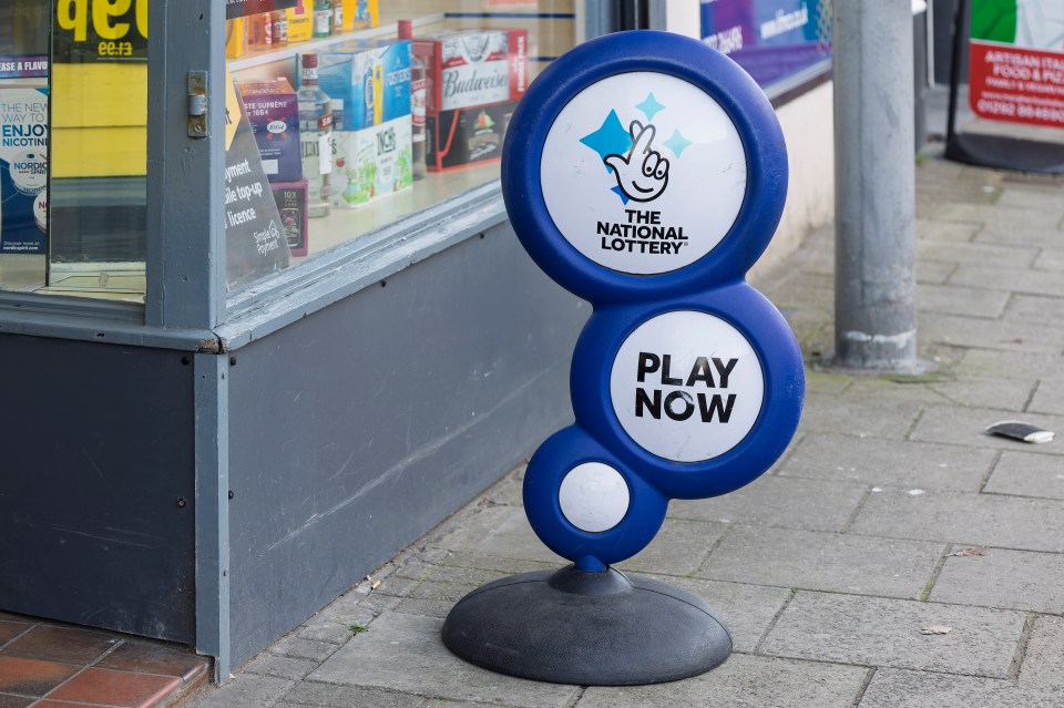A National Lottery promotion sign in Ayr, UK, in front of a shop window.