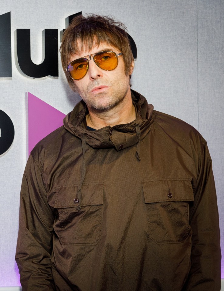 Liam Gallagher wearing a brown jacket and yellow-tinted sunglasses, standing in front of an Absolute Radio sign.