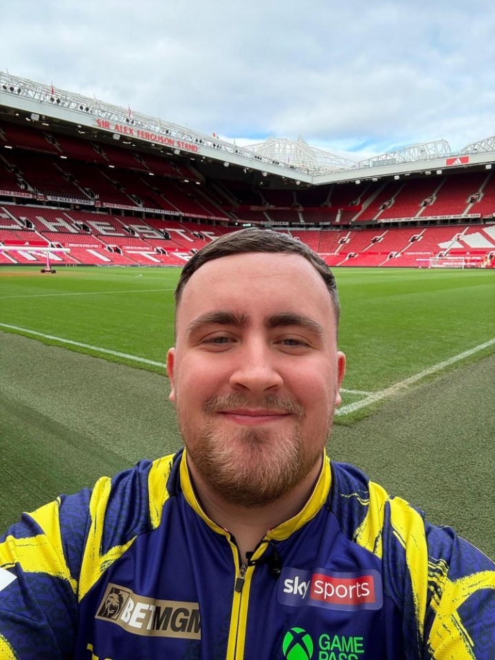 A man in a blue and yellow athletic jacket smiling in front of a large, empty football stadium.