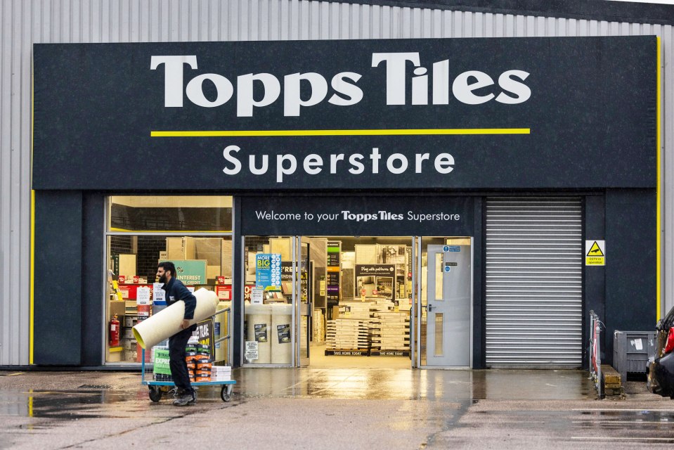 A trade worker exits a Topps Tiles Plc superstore, pushing a trolley laden with materials and carrying a large rolled item.