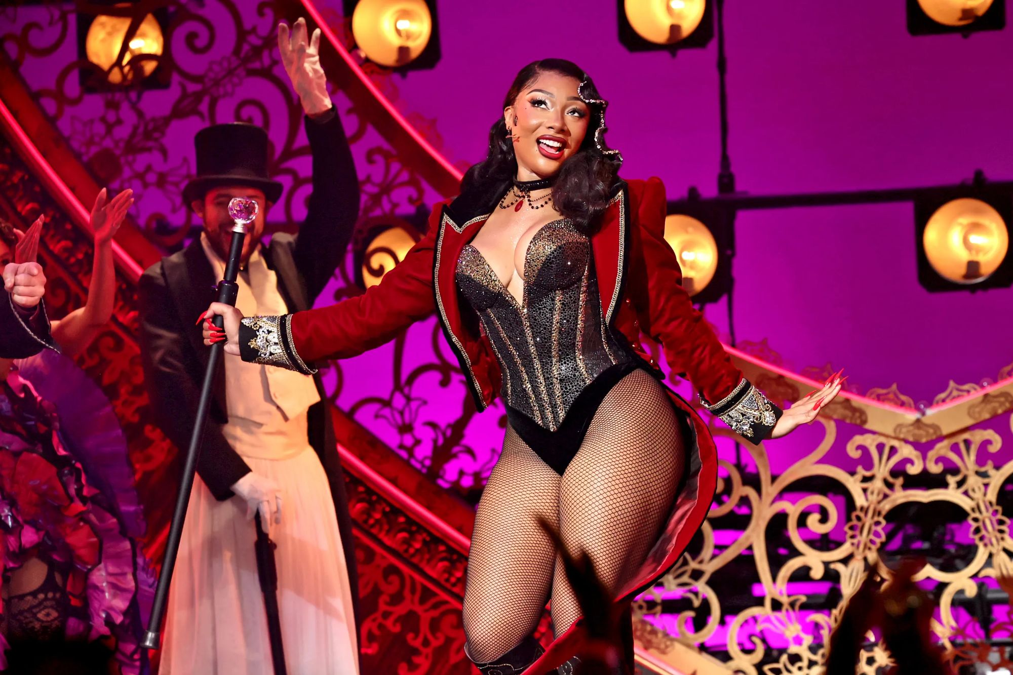 An image collage containing 1 images, Image 1 shows Megan Thee Stallion on stage in a red jacket over a black corset and fishnet tights, with male performers in the background