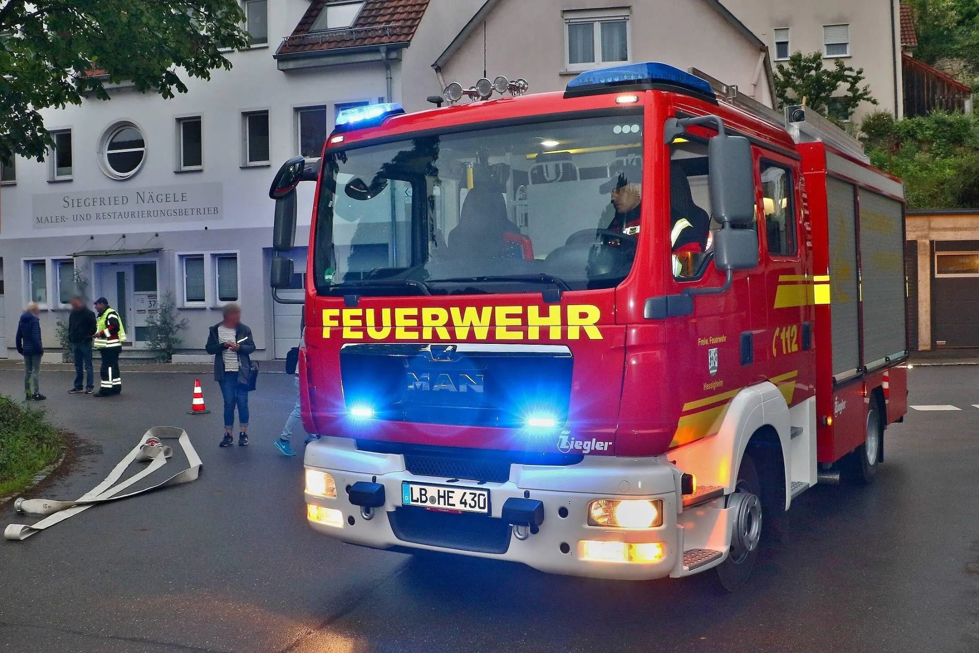 An image collage containing 1 images, Image 1 shows A red fire truck with flashing blue lights and the word "FEUERWEHR" across its front, with people blurred in the background and a white hose on the street in front
