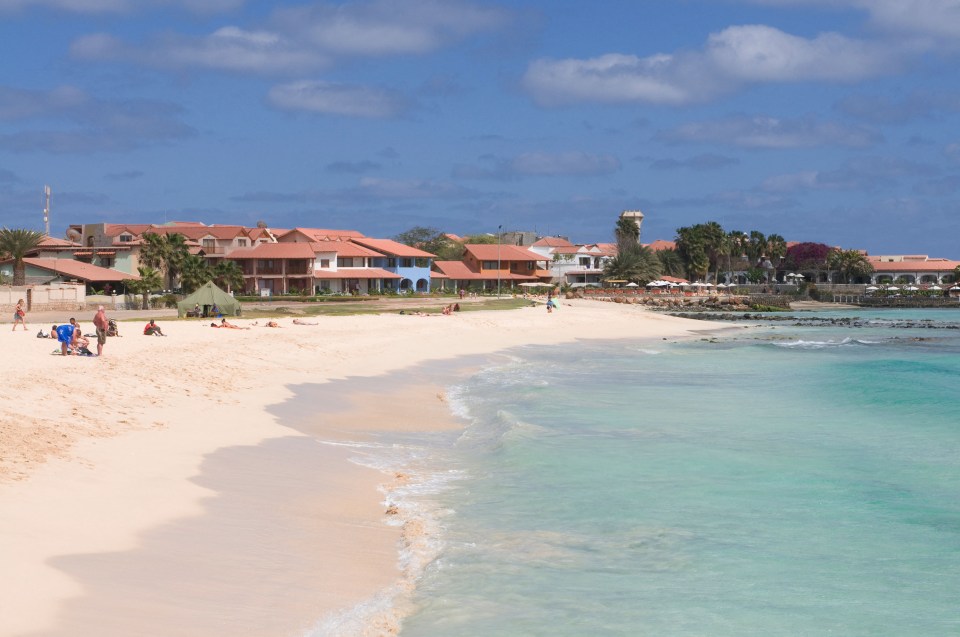 Sandy beach with light blue water and buildings with orange roofs.