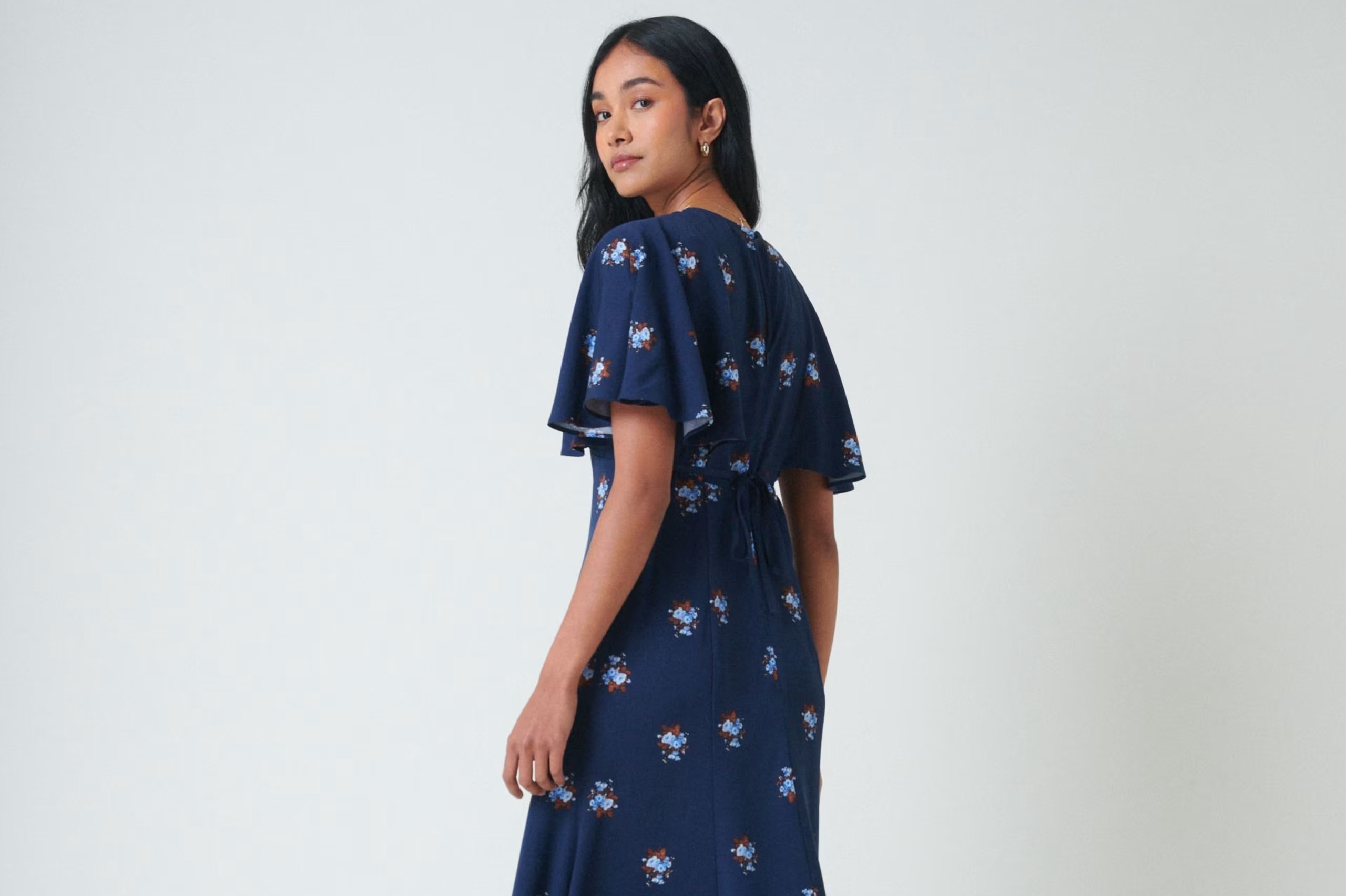 A young woman in a dark blue floral print dress, with her back mostly to the viewer, turns her head to look over her left shoulder.