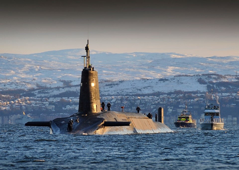 Nuclear submarine HMS Vanguard returns to HM Naval Base Clyde, Faslane, Scotland, accompanied by two smaller patrol boats, with a snow-covered mountainous coastline in the background.