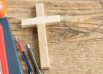 A wooden cross sits on a vintage wooden desk next to some pencils and a stack of notebooks.