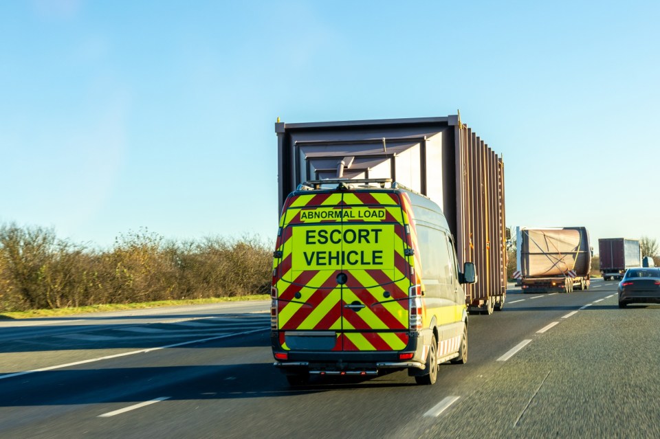 An escort vehicle with "ABNORMAL LOAD ESCORT VEHICLE" written on the back, leading a large transporter carrying a giant silo on a motorway.