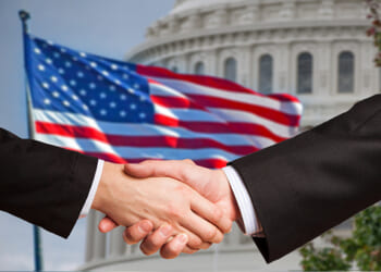 Two men shake hands as the American flag waves behind the United States Capitol rotunda.