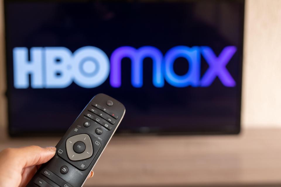 A hand holding a remote control in front of a TV displaying the HBO Max logo.