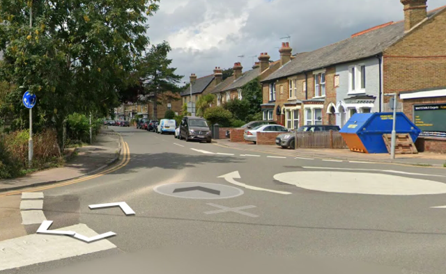 Street view of a road with houses, parked cars, and a blue skip on the right, with a painted roundabout and navigation arrows on the road.