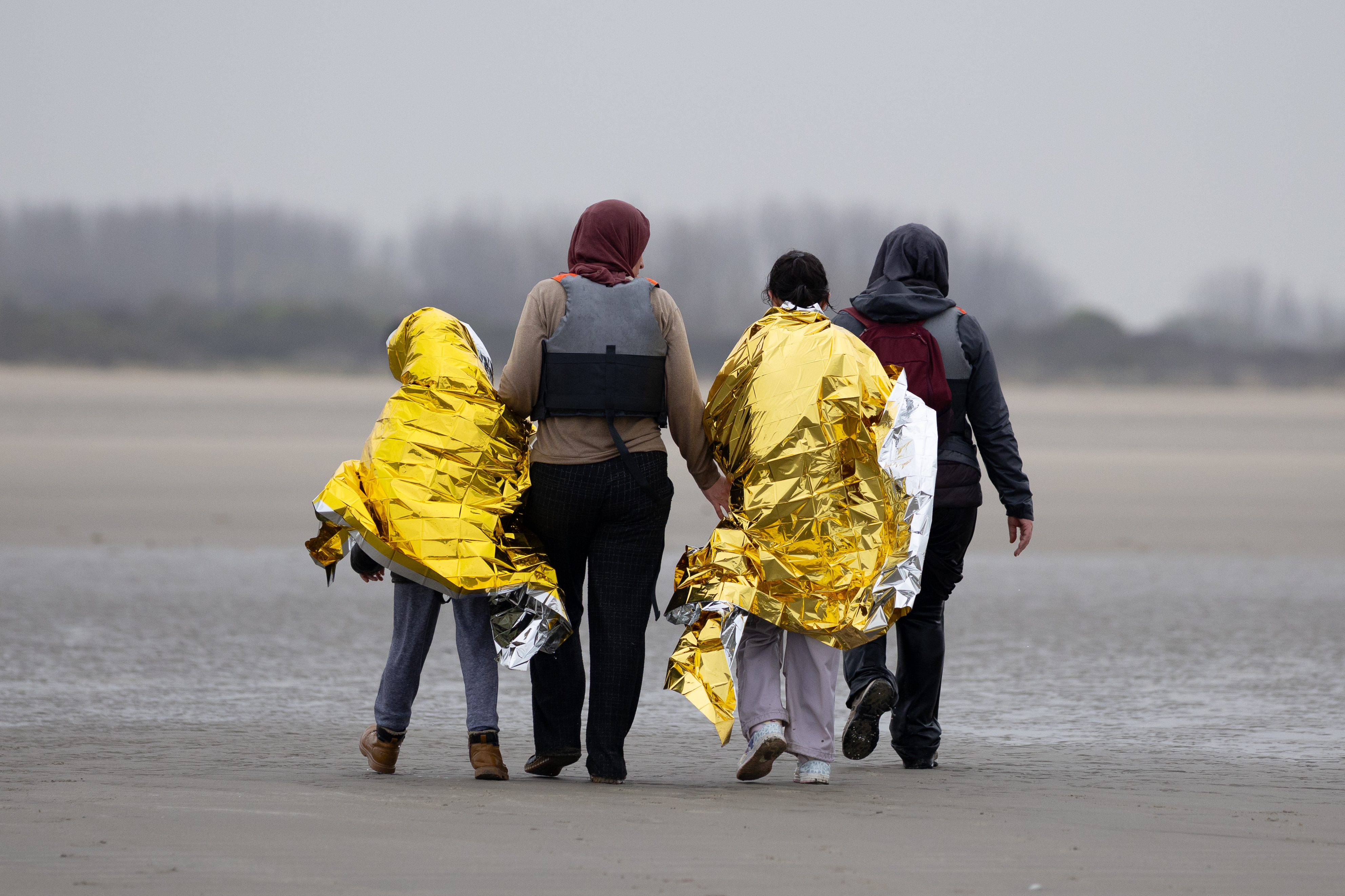 Two children in foil blankets and two women walk up a beach after failing to board a dinghy.
