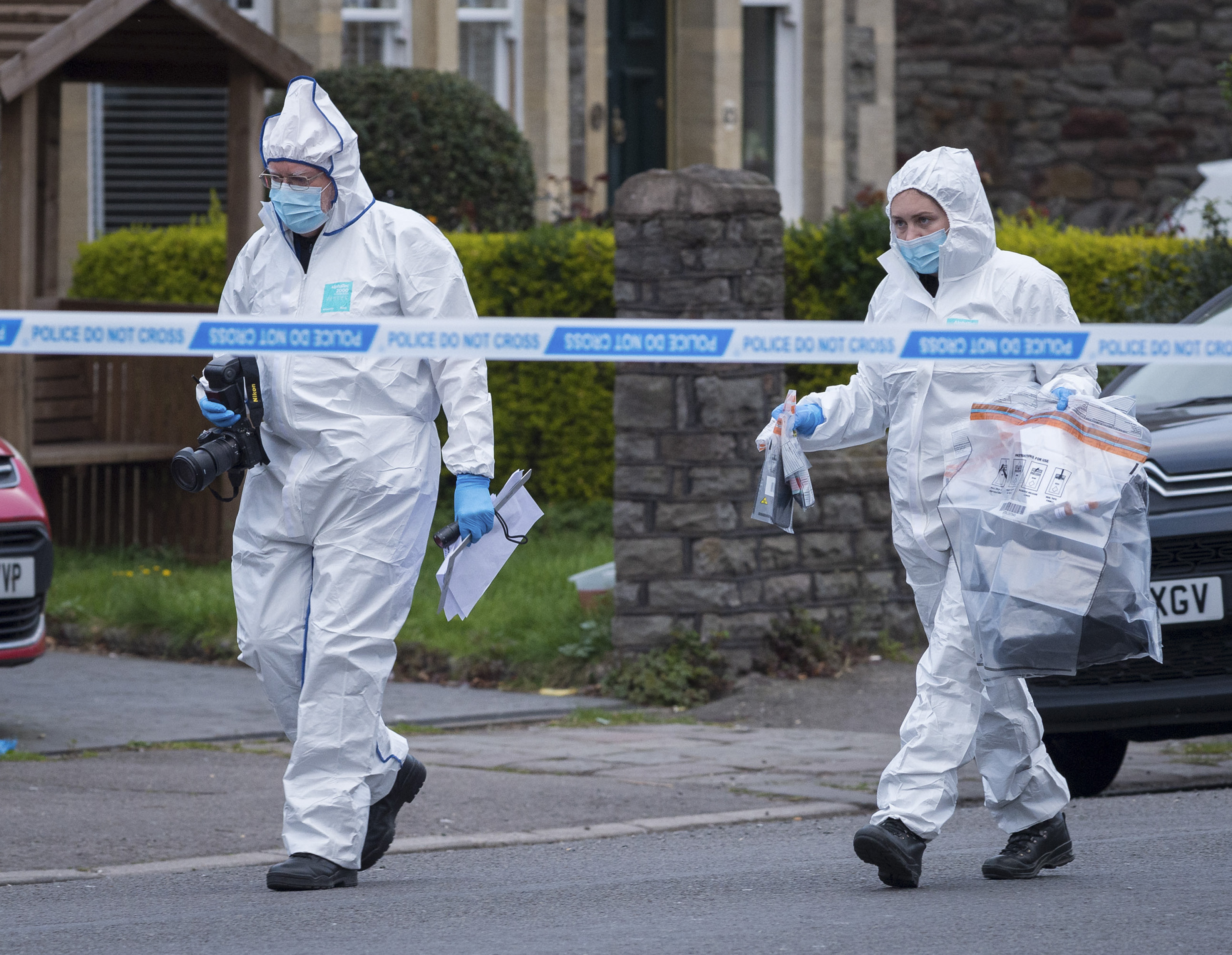 Two forensic officers in white suits and masks are seen behind police tape at a crime scene.