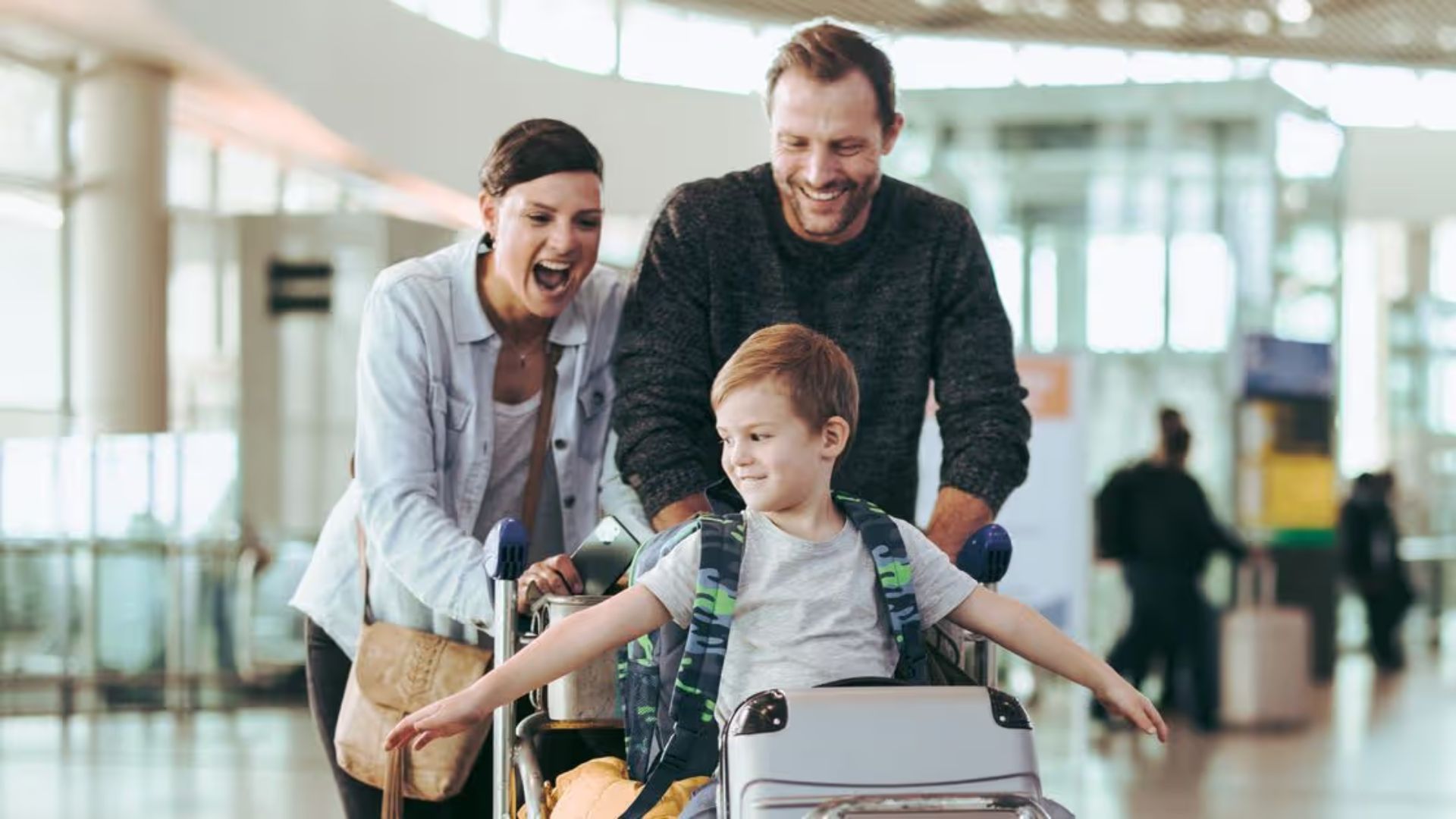A family of three, parents and a young son, smiles as they push luggage through an airport, with the boy sitting on a cart.
