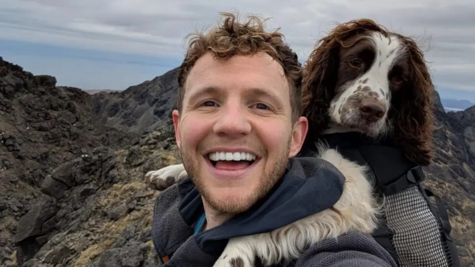 Smiling David Parrish carrying his brown and white dog in a backpack, with a mountainous landscape behind them.