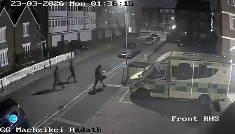 Security camera footage of two people walking on a street next to a parked ambulance at night.