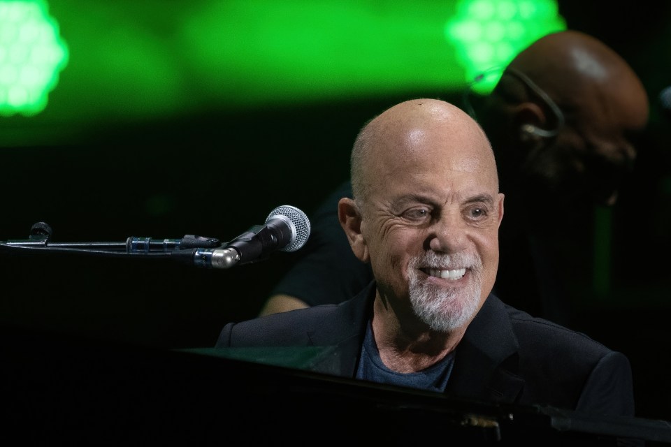 Musician Billy Joel smiles at his piano, with a microphone in front of him and green lights in the background.