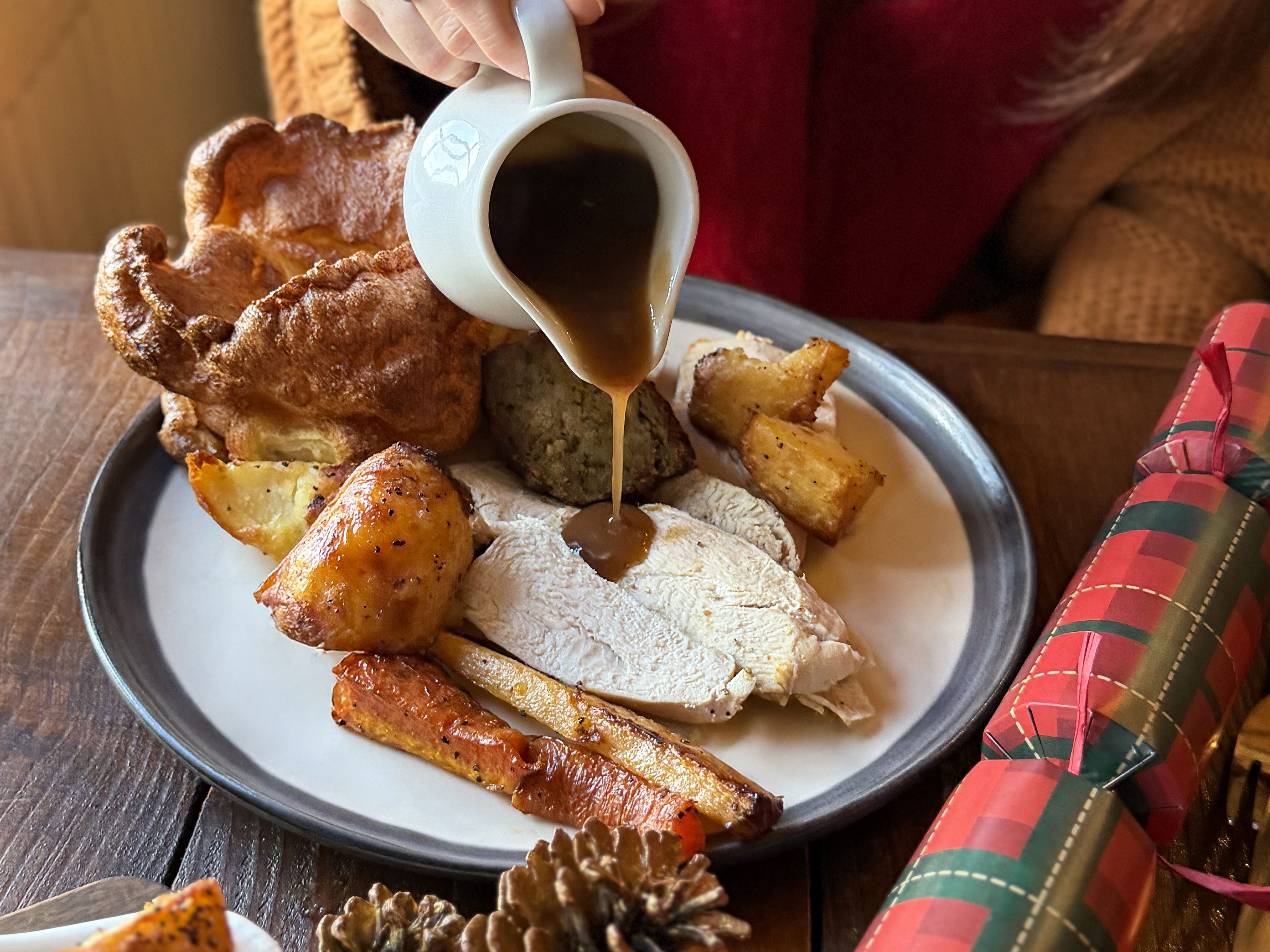 Stock photo showing close-up view of an unrecognisable, young woman in a restaurant eating a Christmas roast dinner comprising of thick slices of roast chicken, Yorkshire pudding, sage and onion bread stuffing, roasted carrots and potatoes, broccoli, cabbage, cauliflower and gravy on white plate. Also pictured are two, tartan patterned, Christmas crackers festive table decorations.