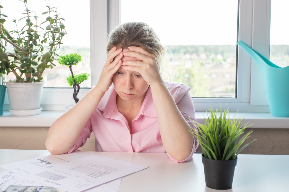A frustrated woman sits at a table with her hands on her head, looking at utility bills.