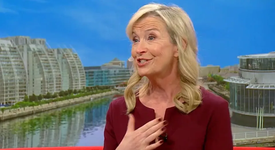 A blonde woman in a red top with her hand on her chest, speaking, with a cityscape background.