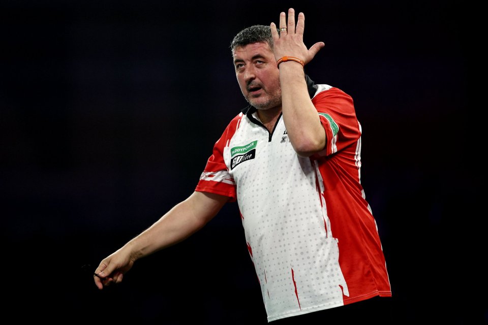 A darts player in a red and white shirt raises a hand to his head, looking frustrated.