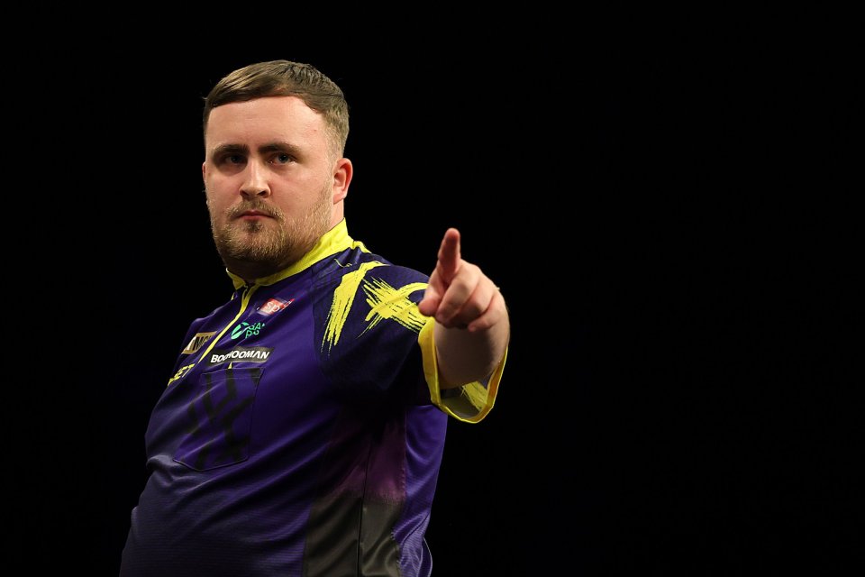 A male darts player in a purple and yellow shirt points his right index finger forward.