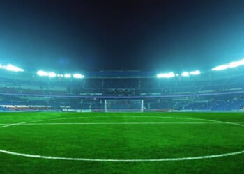 An illuminated soccer stadium at night with a green field and empty stands, ready for an exciting match under bright blue floodlights.