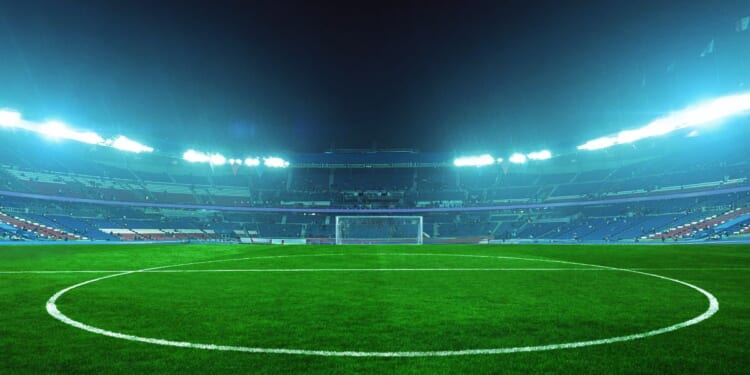 An illuminated soccer stadium at night with a green field and empty stands, ready for an exciting match under bright blue floodlights.